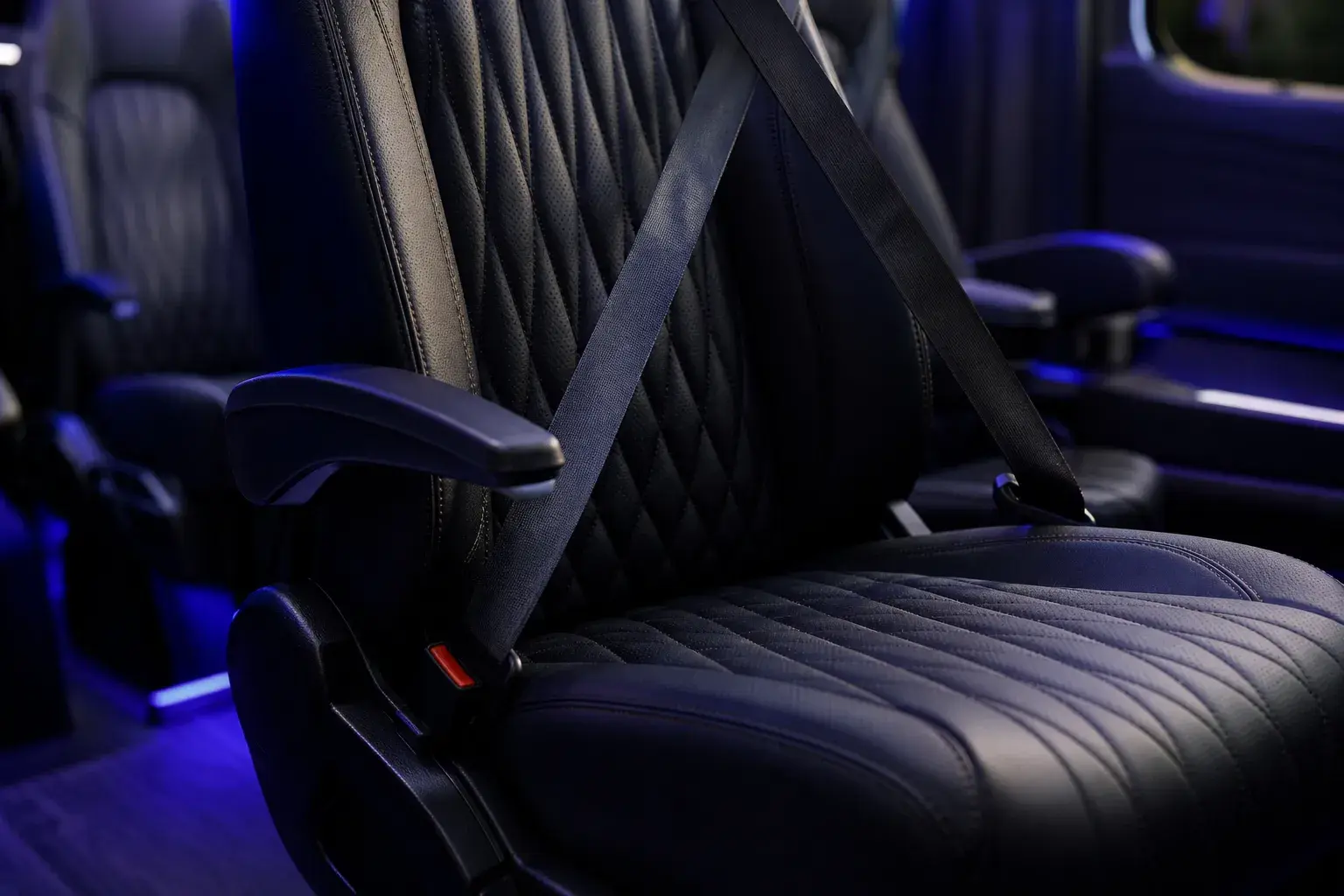 Luxury Sprinter seat detail — black quilted diamond-stitched leather captain's chair with armrest and seatbelt, blue ambient lighting